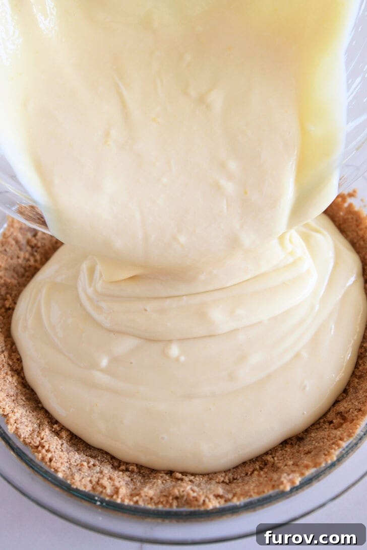 Lemon pie filling getting poured into a graham cracker crust in a glass pie pan.