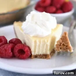 A slice of lemon pie with a graham cracker crust, topped with whipped cream and a bite taken out. Fresh raspberries sit beside the slice.