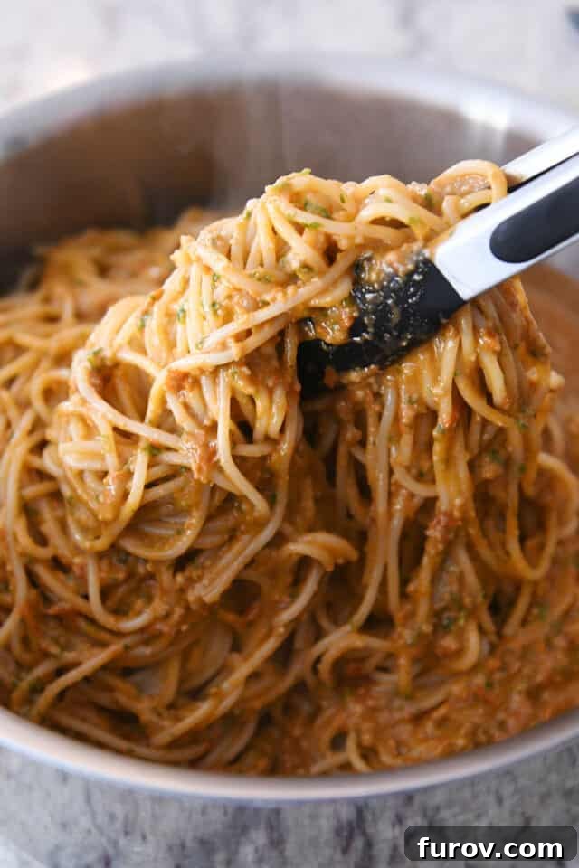Tongs lifting up pasta and pesto sauce in stainless pot.