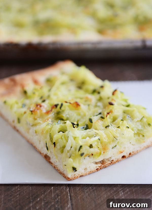 A generous slice of golden zucchini and cheese pizza, with a single bite taken out, resting on a white napkin.