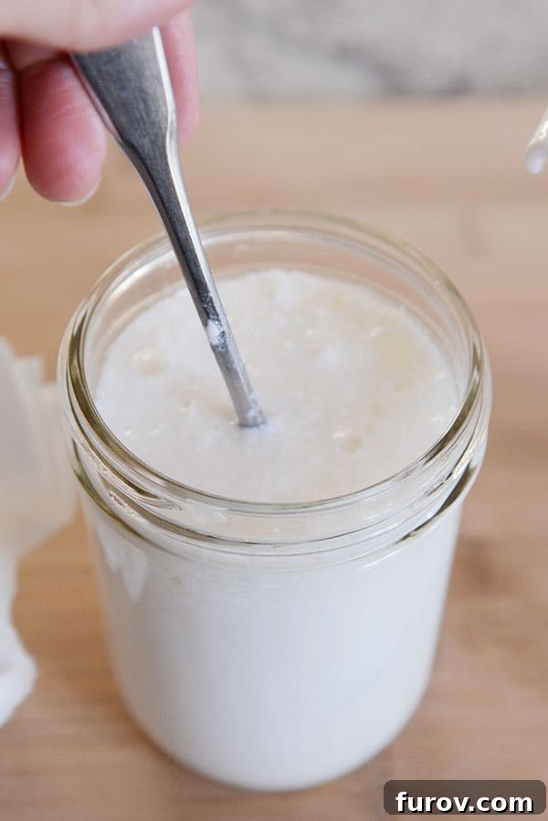 A spoon actively stirring milk kefir and grains inside a mason jar, preparing it for straining.