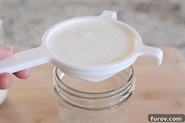 A plastic fine-mesh strainer with kefir grains and thick kefir liquid, straining into a clean mason jar below.