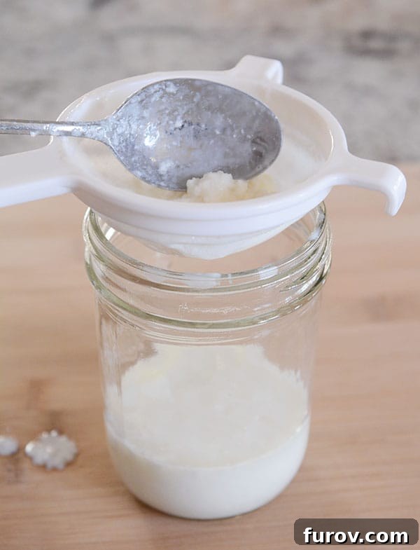 Close-up view of milk kefir grains resting in a plastic strainer positioned over a mason jar, with finished kefir collecting below.