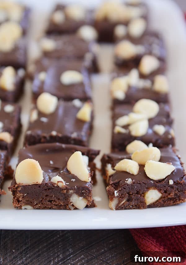 A white platter showcasing beautifully cut chocolate macadamia nut brownies, topped with a glossy fudge and scattered macadamia pieces.