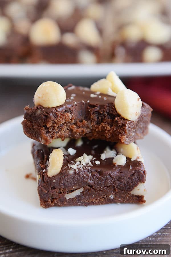 A stack of two luscious chocolate macadamia nut brownies, with a tempting bite taken out of the top one, revealing its soft interior.