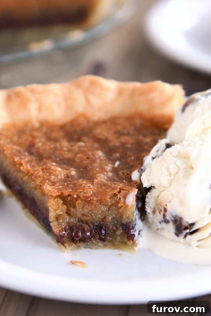 A perfectly baked slice of chocolate chip cookie pie, golden brown and wonderfully gooey, with a single bite taken out. It rests on a pristine white plate, accompanied by a scoop of creamy vanilla ice cream, hinting at the ultimate dessert indulgence.