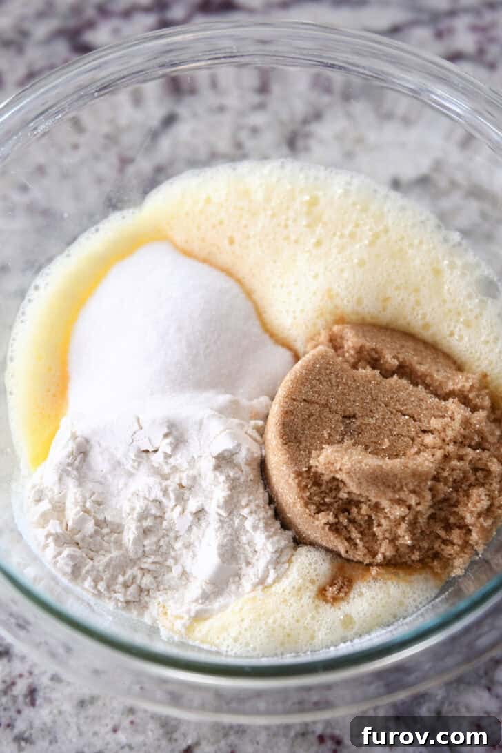 A top-down view of the initial ingredients for the chocolate chip cookie pie filling, including eggs, flour, sugars, salt, and vanilla, neatly arranged in a clear glass mixing bowl, ready to be combined.