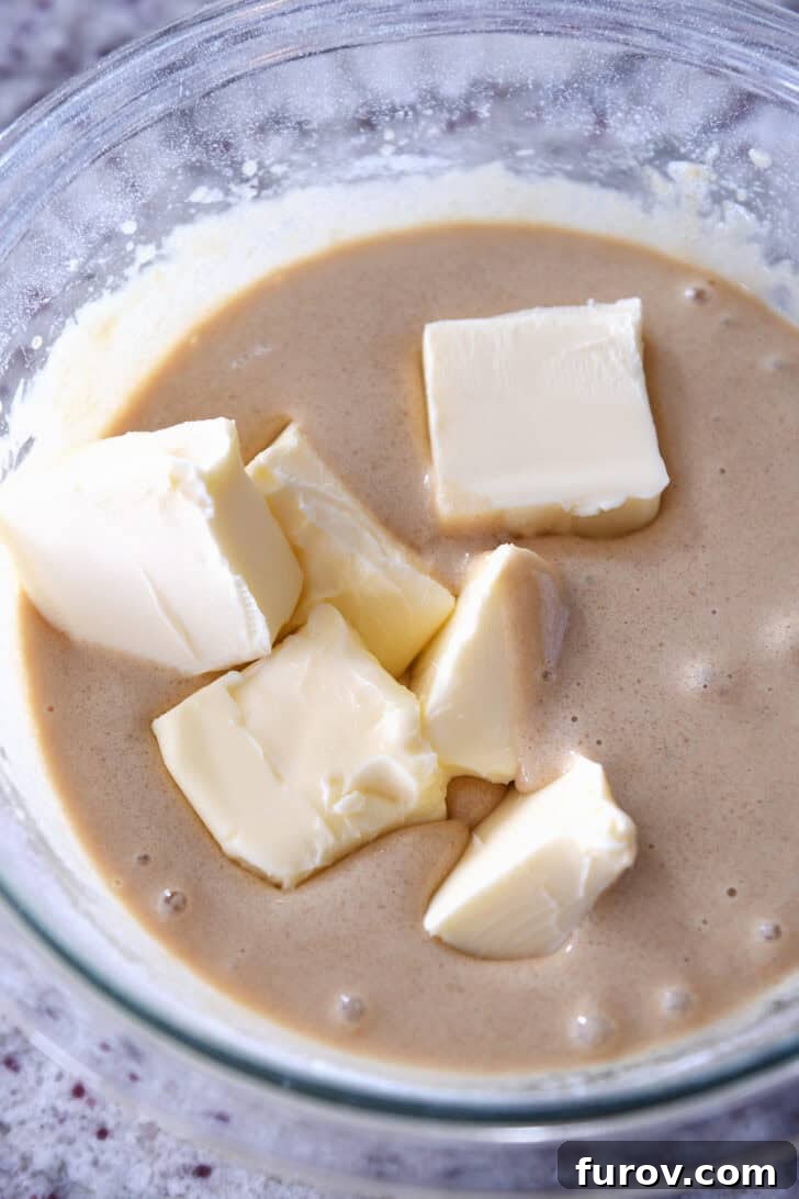 A top-down view of the cookie pie batter in a glass bowl, with softened butter cubes just added, illustrating the step where the rich butter is incorporated into the mixed dry and wet ingredients.
