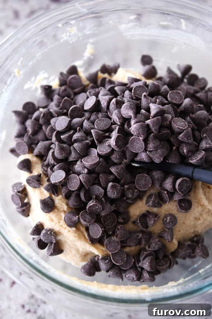 A top-down view of the prepared chocolate chip cookie dough batter in a glass bowl, now studded with an abundance of semisweet chocolate chips on top, indicating it's ready for baking.