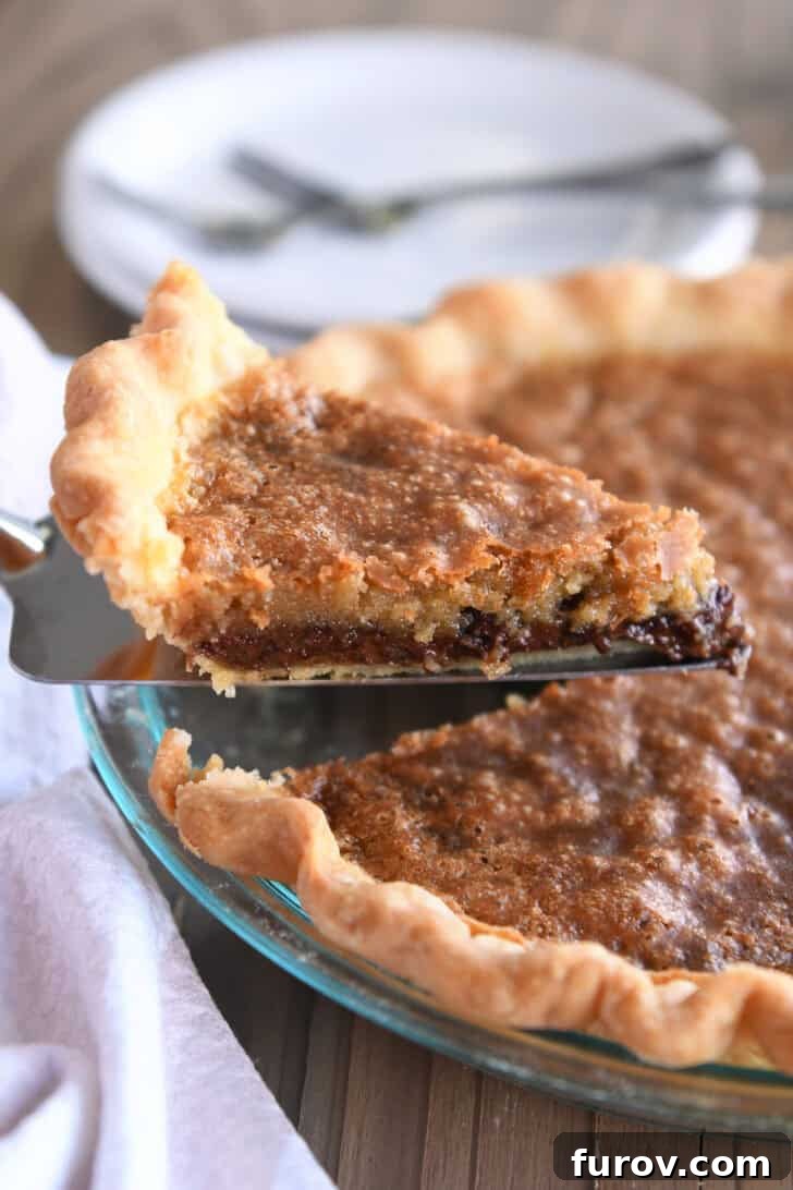 A glistening metal spatula expertly slides underneath a perfectly baked slice of chocolate chip cookie pie, carefully lifting it away from the rest of the pie, highlighting its rich, golden-brown crust and tempting gooey center.