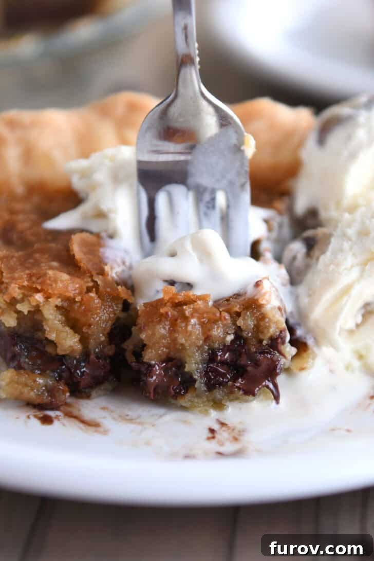 A pristine white plate holds a generous slice of golden-brown chocolate chip cookie pie, its gooey center visible. A fork is poised, having just taken a perfect bite, showing the rich, melted chocolate within.