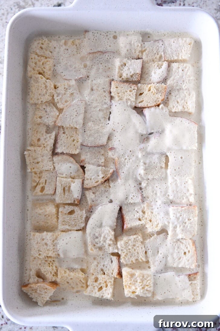Top-down view of a white ceramic baking dish with the egg and half-and-half custard mixture being poured over the bread chunks, ensuring even saturation.