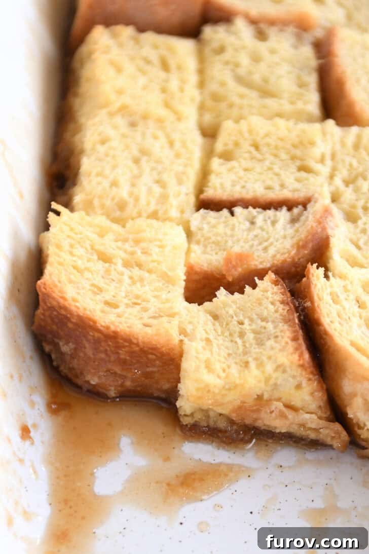 Top-down view of a white ceramic baking dish featuring egg-coated bread, with a section scooped out, revealing the custardy interior of the baked French toast.