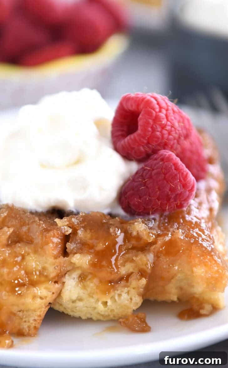 A white plate holds a slice of baked creme brûlée French toast, beautifully topped with a dollop of whipped cream and vibrant fresh raspberries.