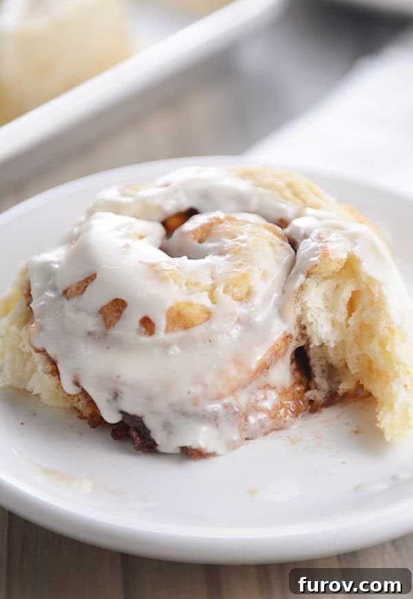 A single frosted buttermilk biscuit cinnamon roll on a white plate, with a piece gently pulled away to show its flaky interior.
