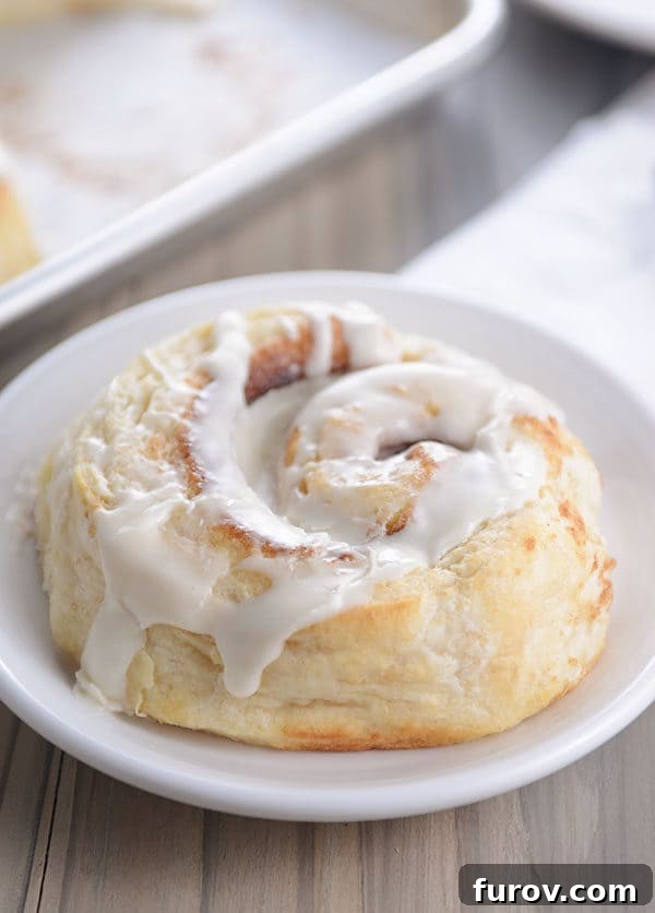 A single, perfectly round buttermilk biscuit cinnamon roll, generously covered in white icing, resting on a small white plate.