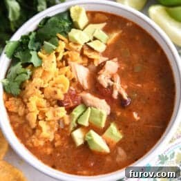 Top down view of a white bowl full of chicken tortilla soup topped with diced avocados, crushed chips, and chopped cilantro.
