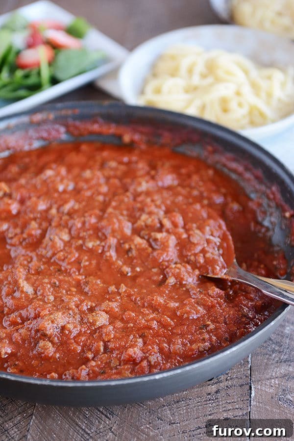Speedy Homemade Weeknight Marinara 3 A large skillet brimming with rich, meaty red spaghetti sauce, with a fresh green salad and cooked spaghetti noodles blurred in the background, suggesting a complete meal.