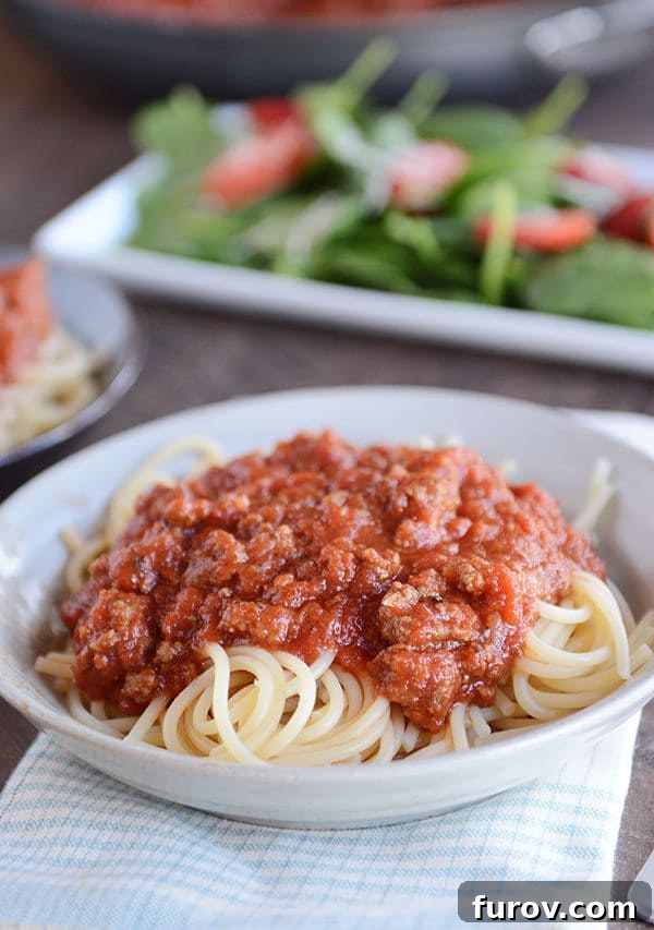 Speedy Homemade Weeknight Marinara 4 A white bowl of spaghetti noodles generously covered with meaty, red spaghetti sauce, with a fresh green salad visible in the background, making for a balanced meal.