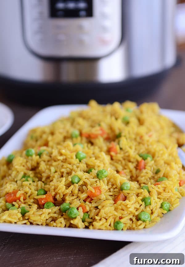 A white plate full of vibrant, yellow-tinted Indian vegetable rice, generously sprinkled with bright green peas and tender carrots, with an Instant Pot softly blurred in the background.