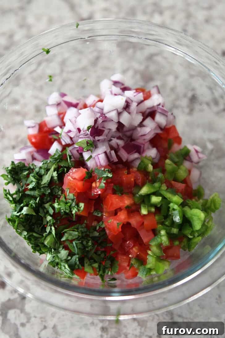 A glass bowl filled with vibrant, freshly chopped ingredients for pico de gallo: tomatoes, red onion, cilantro, and jalapeños.
