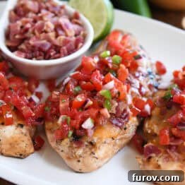 Grilled chicken breasts topped with pico de gallo on a white plate with a bowl of chopped bacon and a half of a lime in the background.