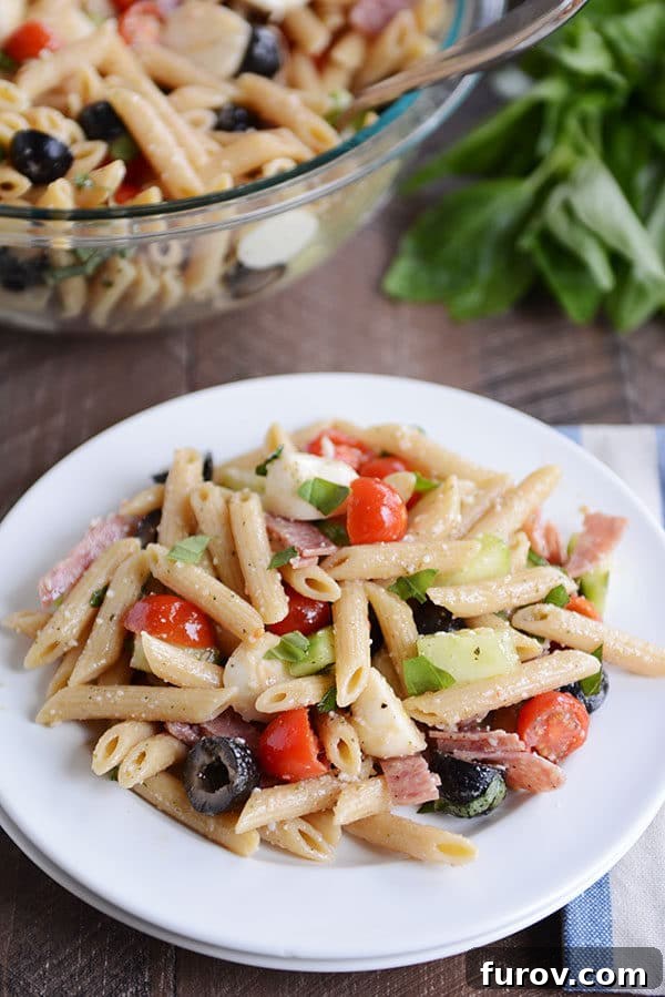 A white plate of penne pasta salad with olives, tomatoes, onions, mozzarella, and basil, garnished with fresh herbs.