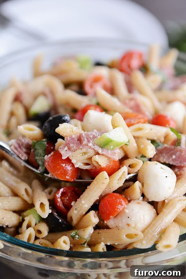A spoon taking a scoop out of a glass bowl of Italian pasta salad, highlighting the various ingredients.