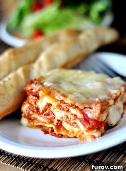 A generous slice of classic Italian lasagna, rich with meat, creamy white sauce, and bubbling cheese, served on a pristine white plate alongside golden breadsticks, promising an exquisite meal.