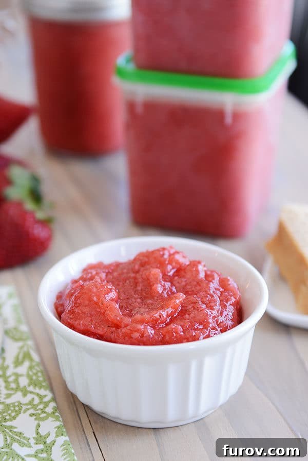 A small white ramekin of fresh strawberry jam with clear plastic containers of more jam behind it. Highlighted by natural light, the jam's vibrant red color promises fresh, sweet flavor.