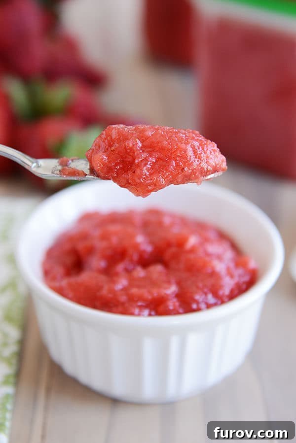A silver knife carefully scoops a dollop of vibrant, fresh strawberry jam from a small white ramekin, highlighting its tempting texture.