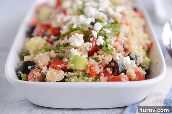 A white dish full of cooked quinoa with chopped cucumbers, olives, and tomatoes and crumbled feta on top.