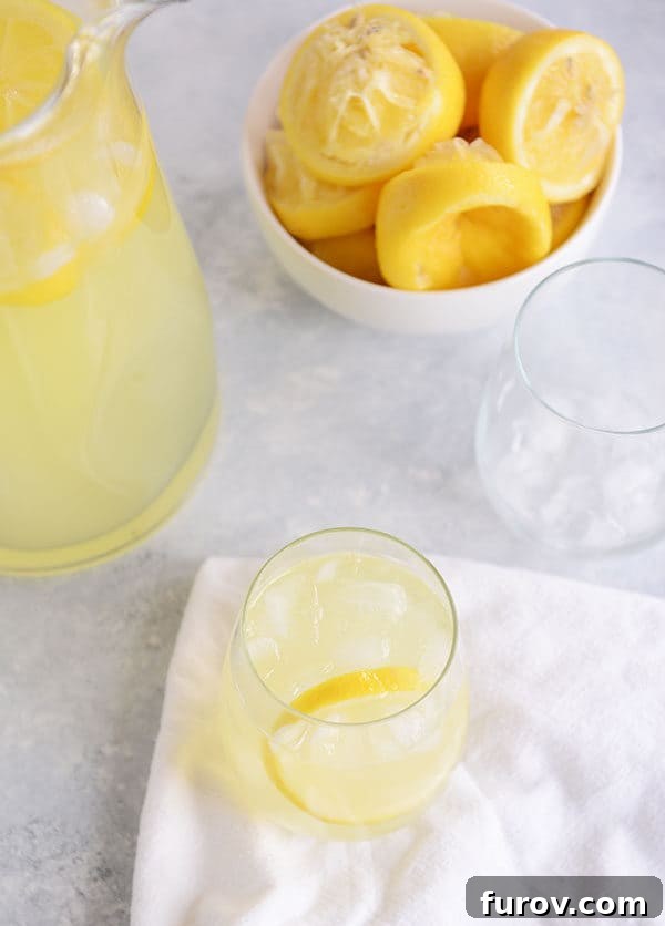 Top view of a glass of lemonade with ice and a bowl of lemons and a pitcher of lemonade beside it.