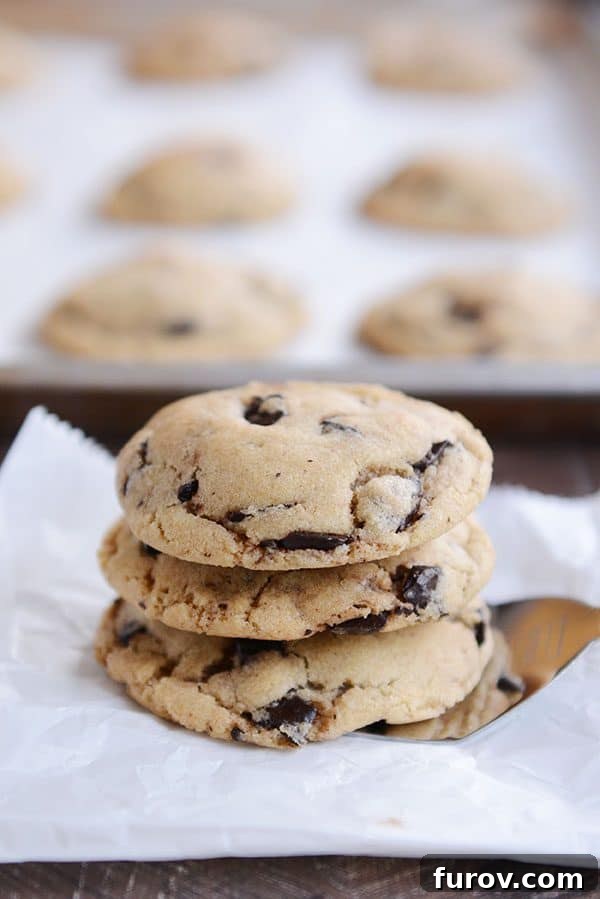 Three perfectly baked chocolate chip cookies stacked on a metal spatula with a tray of more cookies cooling in the background.
