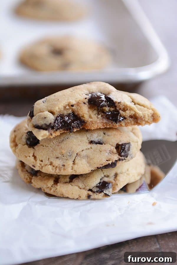 Freshly baked chocolate chip cookies stacked on a metal spatula with the top cookie split in half to show its gooey center.