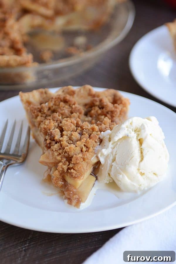 No-Fuss Apple Crumble Pie 2 A perfectly baked slice of apple crumble pie resting on a white plate, accompanied by a generous scoop of creamy vanilla ice cream. The golden-brown streusel topping is visibly crisp and inviting.