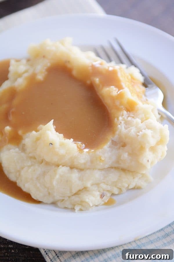 Hands-Off Creamy Slow Cooker Mashed Potatoes 3 A white plate with a large serving of mashed potatoes covered in rich brown gravy.