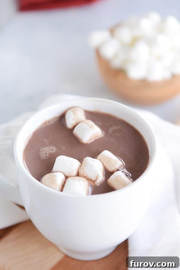 A white mug of steaming hot chocolate and marshmallows floating on the top with a bowl of marshmallows in the background.