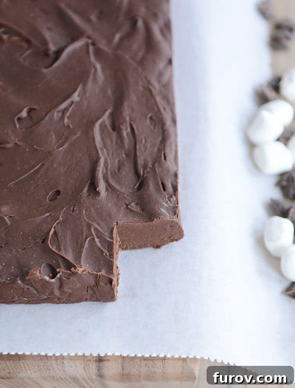 A brick of fudge on white parchment paper, with one square cut out in the corner, and marshmallows and chocolate chips sprinkled on the side of the parchment paper. 
