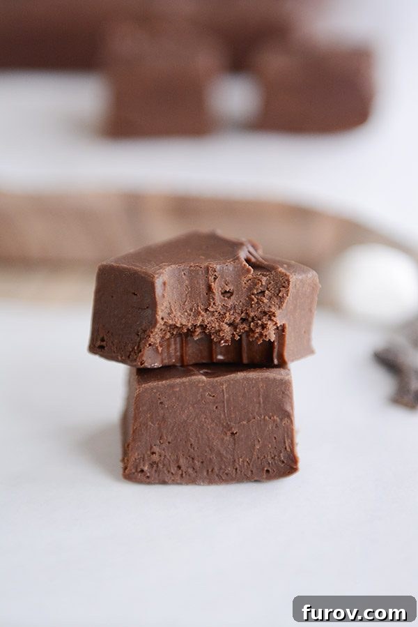 Two squares of fudge stacked on top of each other. The top one has a bite taken out of it. In the background there are more squares of fudge. 