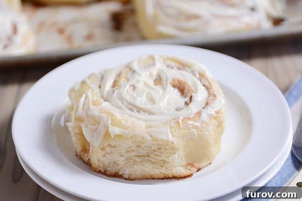 A beautifully frosted cinnamon roll on a white plate, glistening with sweet icing, perfectly ready to be enjoyed.