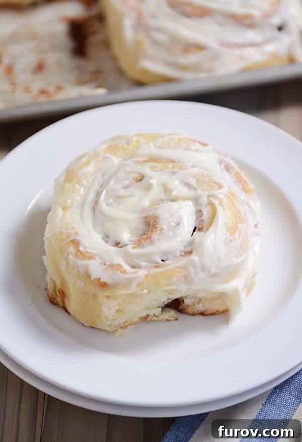 A close-up view of a single frosted cinnamon roll on a white plate, highlighting its soft texture and abundant icing.