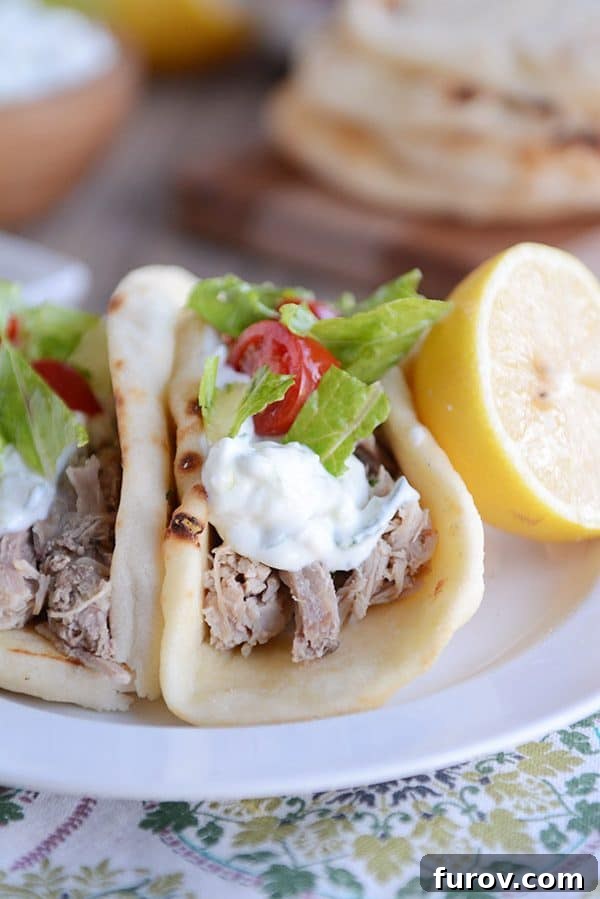 Fall-Apart Tender Greek Pork Instant Pot or Slow Cooker 2 A white plate of two Greek pork tacos topped with lettuce and tomatoes with a half lemon on the side.