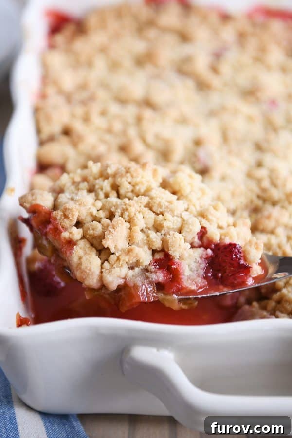A beautiful, golden-brown strawberry rhubarb crumble, freshly baked in a white ceramic pan, with a serving spoon taking out a piece.