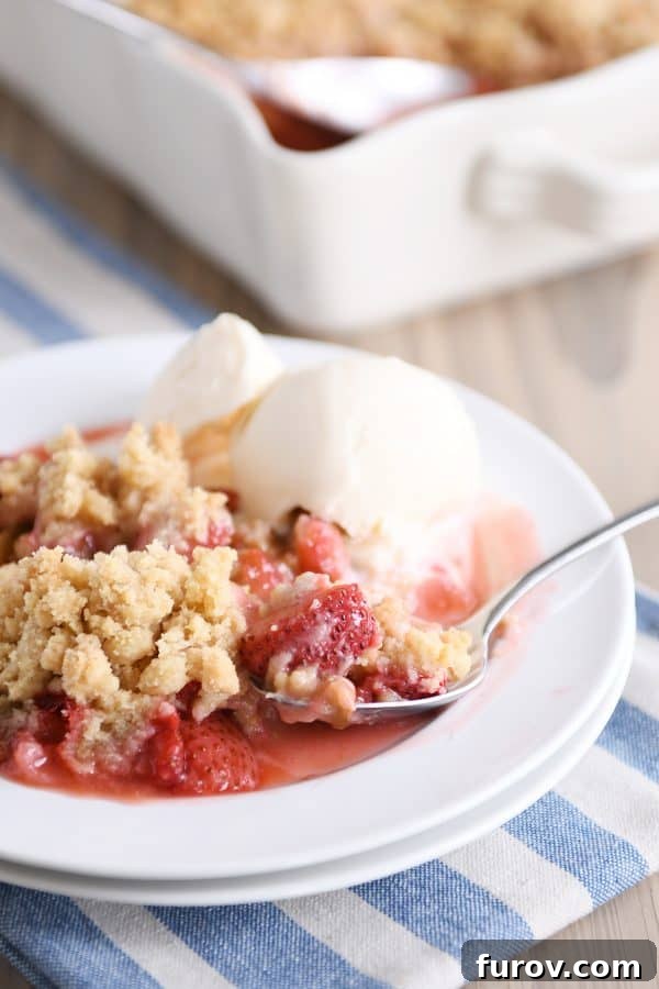 A white plate with a warm scoop of strawberry rhubarb crumble, perfectly paired with a melting scoop of creamy vanilla ice cream.