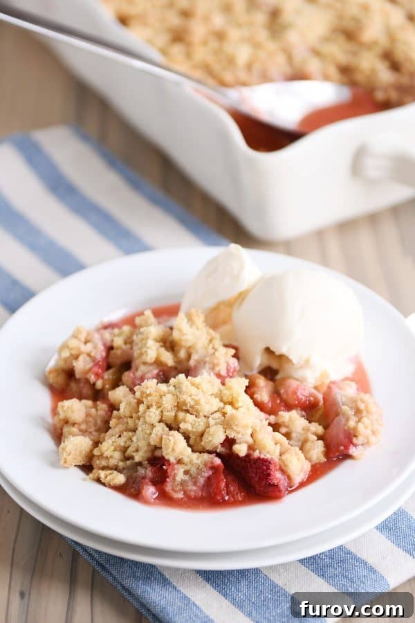 An inviting image of a serving of strawberry rhubarb crumble on a white plate, topped with a scoop of melting vanilla ice cream.