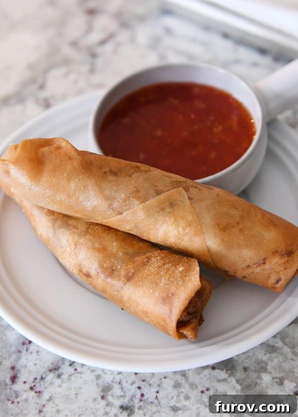 Marys Signature Egg Rolls 2 Two golden-brown, crispy egg rolls served alongside a small bowl of vibrant red dipping sauce on a pristine white plate, promising a delicious culinary experience.