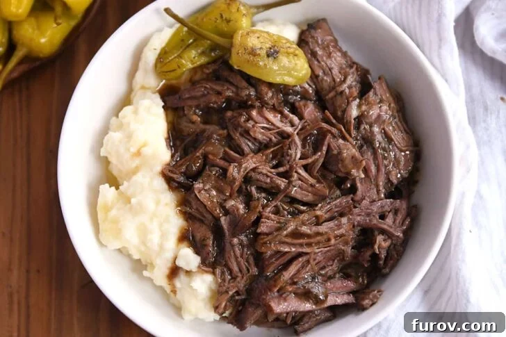 White shallow bowl with mashed potatoes, two pepperoncini, and shredded Mississippi Pot Roast beef.