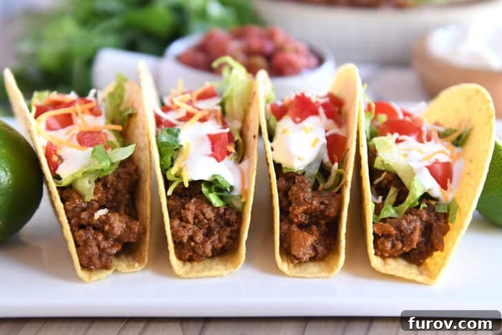 Four crispy-shelled ground beef tacos filled with toppings on a white platter, ready to be served.