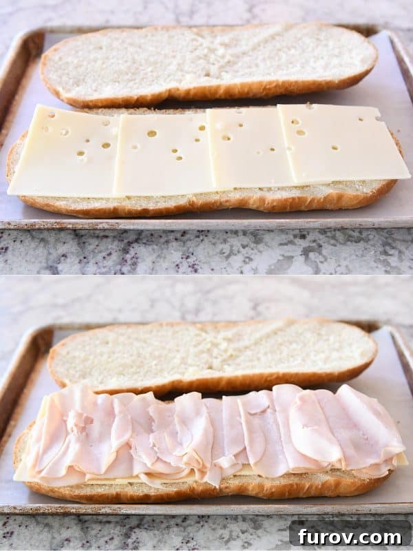 Layers of deli meat and cheese being placed onto the bottom half of a French bread loaf for a sheet pan panini.